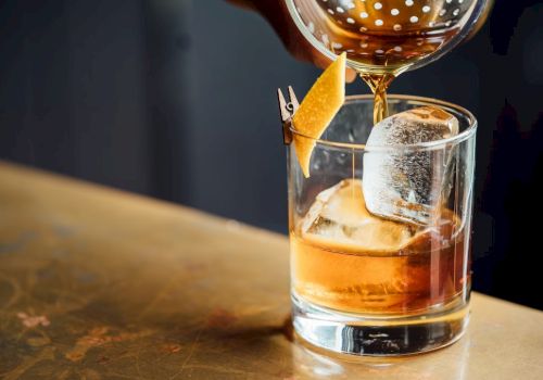 Pouring a drink: a short glass with whiskey on ice, a lemon twist garnish, and a cocktail being poured from a bottle above.