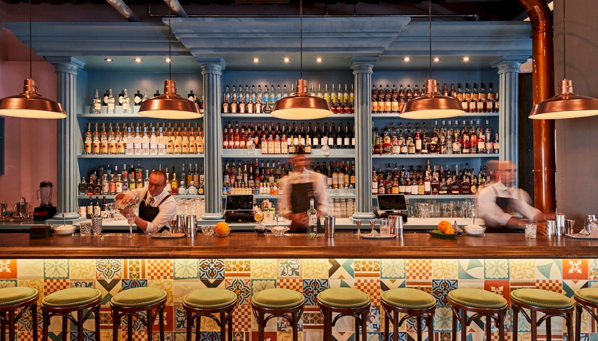 A stylish bar scene with a long counter, colorful tiled front, and shelves full of bottles in the blue back wall.