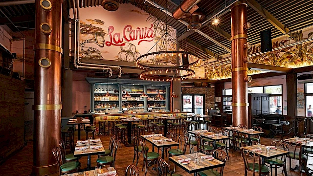 A lively restaurant interior with wooden tables and green chairs, metal pillars, warm lighting, a mural saying &ldquo;La Cantina,&rdquo; and a bar area in the back.