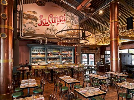 Inside a vibrant La Cantina, rustic tables and green chairs fill a warm, industrial-style dining hall with copper columns and mural art.