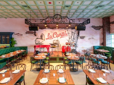 A vintage-style restaurant interior with wooden tables set for dining, green cushioned seating, brick walls, and a mural that reads &ldquo;La Cantina.&rdquo;