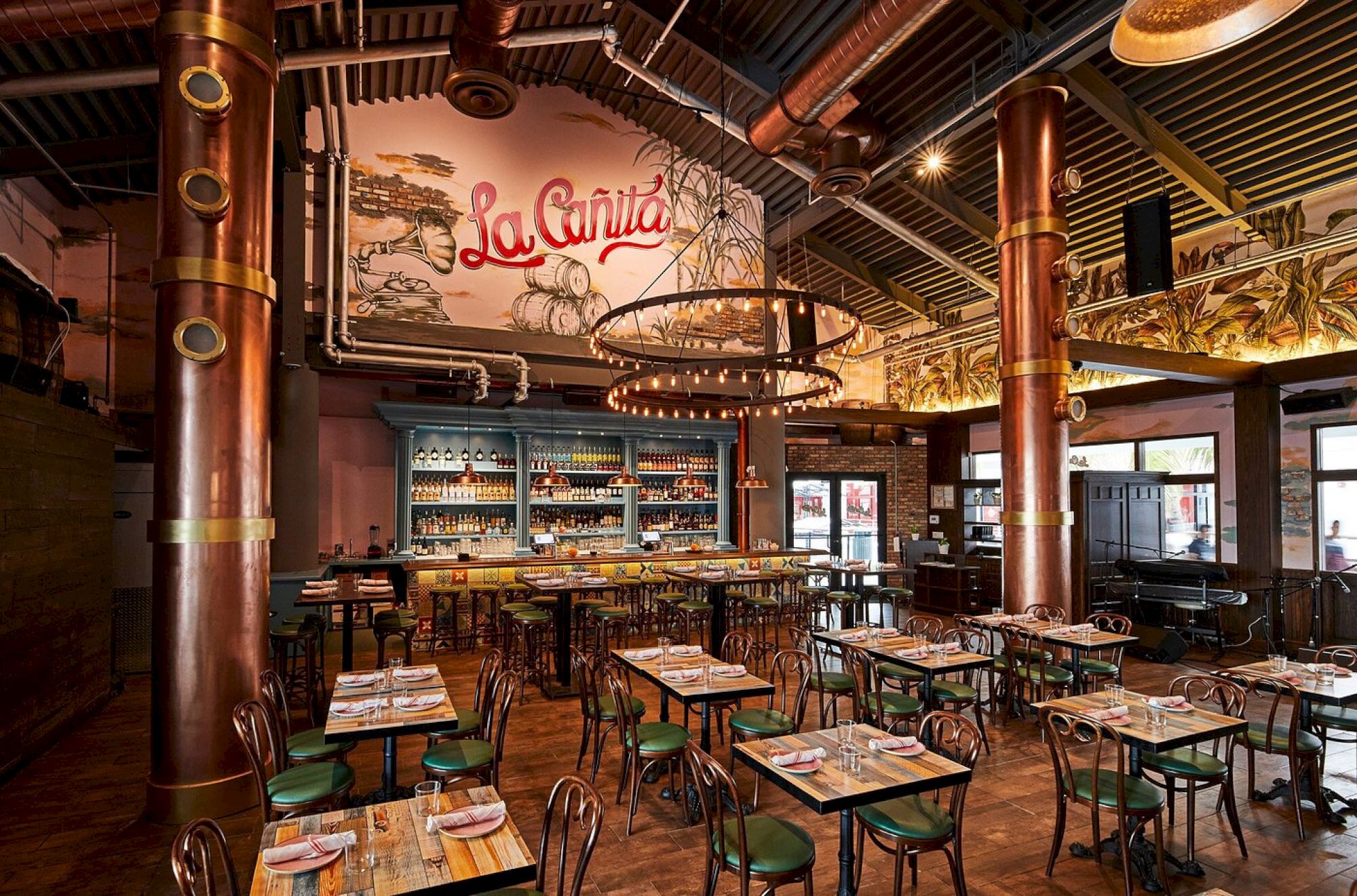 A stylish restaurant with rustic-industrial decor, copper columns, wooden tables, metal chairs, a long bar, and a mural reading &ldquo;La Vida&rdquo; on the wall.