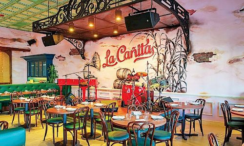 A colorful cafe interior with vintage metal chairs, teal accents, rustic decor, and a sign reading &ldquo;La Ca&ntilde;ita&rdquo; on a painted wall.