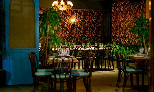 A cozy dining room with tropical-themed wallpaper, green plants, teal chairs, and dim lighting over neatly set tables.