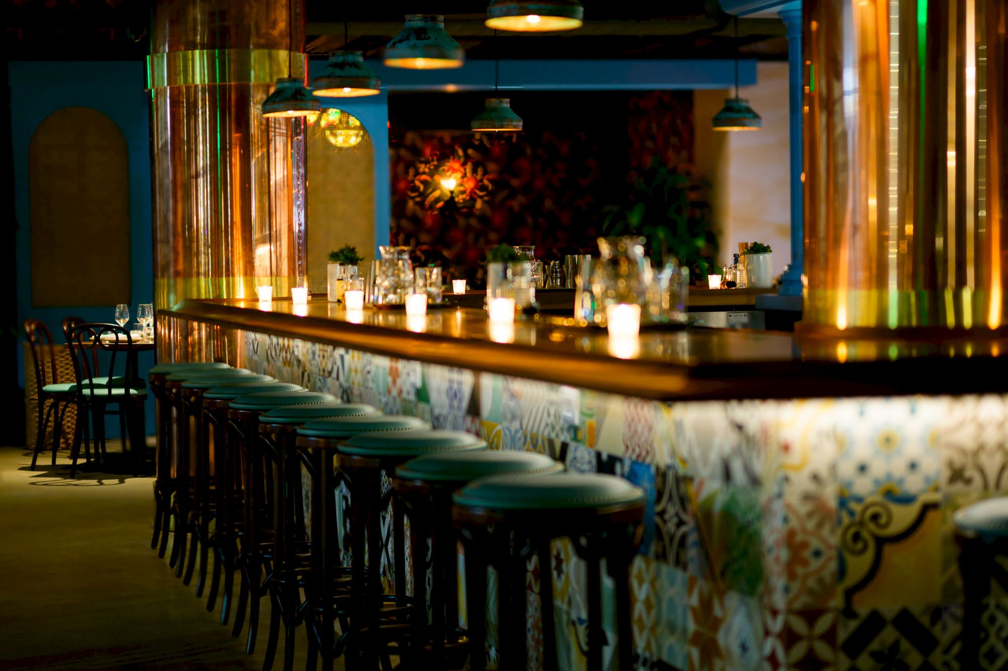 A long bar lined with teal stools, warm candlelight, colorful tiled counter, and blue-green walls in a cozy, stylish lounge.