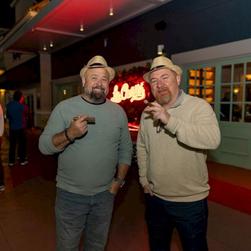 Two smiling men wearing matching beige hats pose with drinks at a lively bar, neon signage glowing behind them.