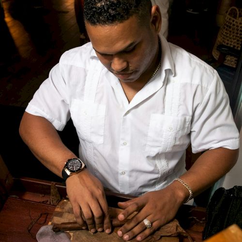 A man in a white shirt is carving or arranging pieces on a wooden board at a table, focused on his hands, with a watch and ring visible, in a dim setting.