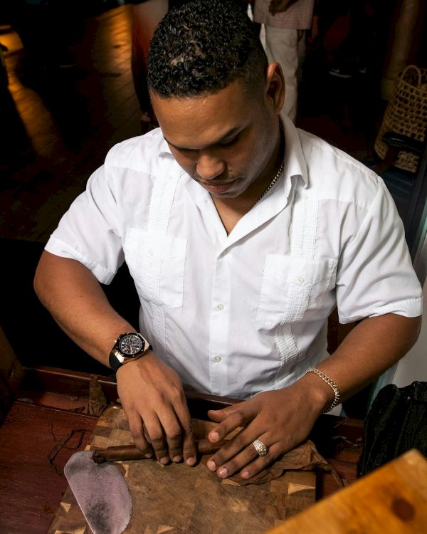 A man in a white shirt is carving or arranging pieces on a wooden board at a table, focused on his hands, with a watch and ring visible, in a dim setting.