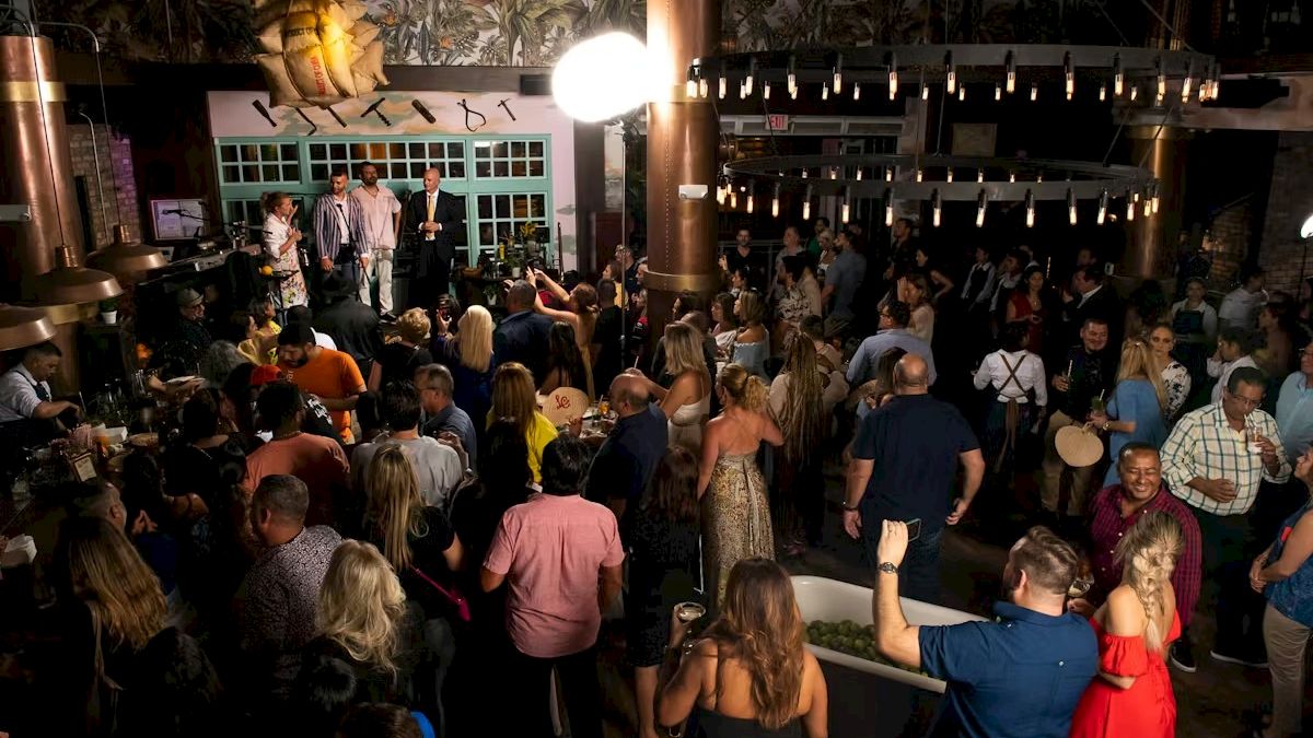 A crowded nightclub or party scene with people dancing and mingling, string lights, a stage or DJ area, and warm, dim lighting.