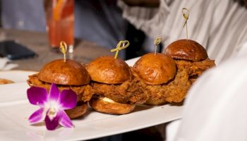 Five mini fried chicken sliders on a white plate, with decorative toothpicks and a purple orchid garnish.