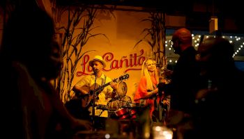 A small live band performs on a dimly lit stage with a guitarist and a female singer, under a warm &ldquo;La Canita&rdquo; sign, as silhouettes crowd nearby.