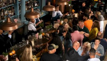 A crowded bar or pub with many patrons standing, chatting, and drinking; bartenders mix drinks under warm pendant lights.