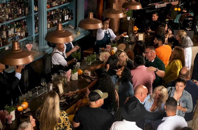 A crowded bar or pub with many patrons standing, chatting, and drinking; bartenders mix drinks under warm pendant lights.