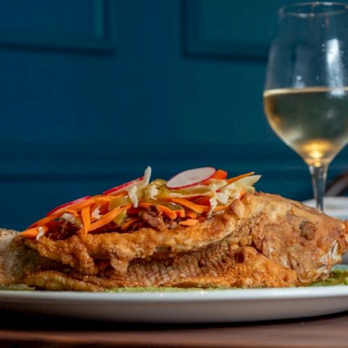 A large sandwich or open-faced dish topped with shredded vegetables on a plate, with a glass of white wine in the background.