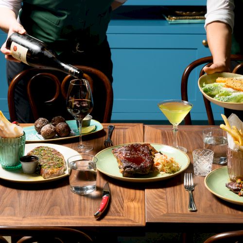 Two waiters serve a fancy table with assorted dishes and drinks; a carafe pours red wine while plates feature steak, fries, and desserts.