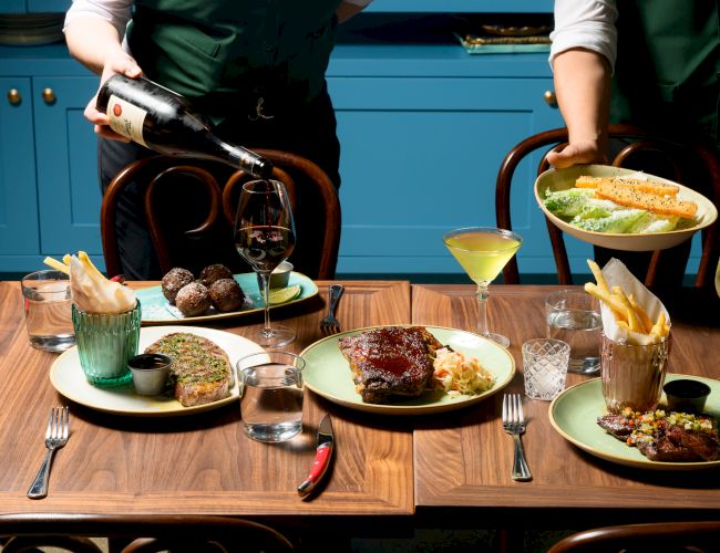 Two waiters serve a fancy table with assorted dishes and drinks; a carafe pours red wine while plates feature steak, fries, and desserts.