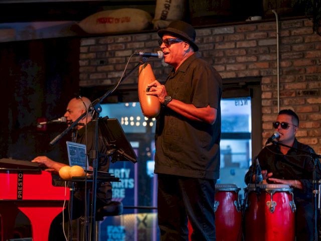 A live band performs on a small stage: a man with a hat sings into a microphone, keyboardist on the left, and percussionists in the back, brick wall backdrop.