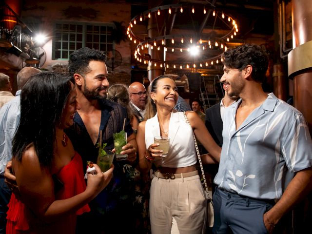 A lively party scene with four friends laughing and chatting, drinks in hand, under warm string lights in a crowded venue.