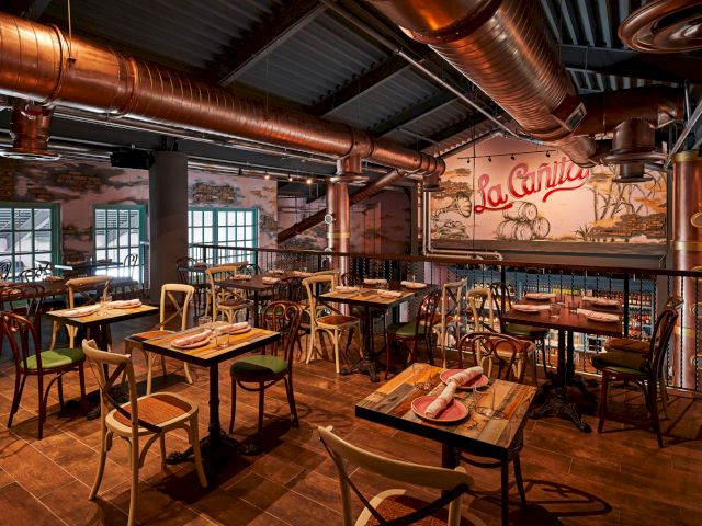 Industrial chic restaurant interior with wooden tables, metal chairs, exposed copper pipes, brick walls, and a mural reading &ldquo;La &hellip;&rdquo; in warm lighting.