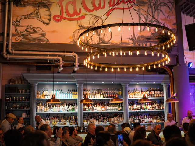 A lively bar scene with people dining at tables, a large circular chandelier, and a mural that says &ldquo;La Cantina&rdquo; above a well-stocked bottles shelves.