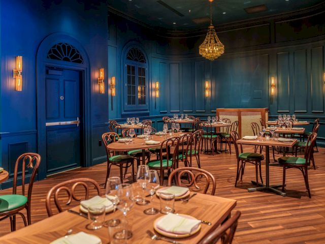 A stylish, blue-walled restaurant with wooden tables, green chairs, warm lighting, and a chandelier, set for a fine dining experience.
