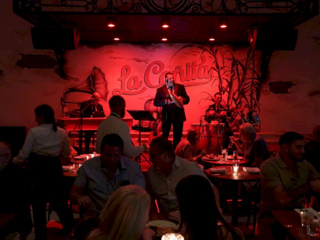A live guitarist performs on a small stage in a dim, red-lit restaurant, surrounded by seated patrons and intimate candlelight.