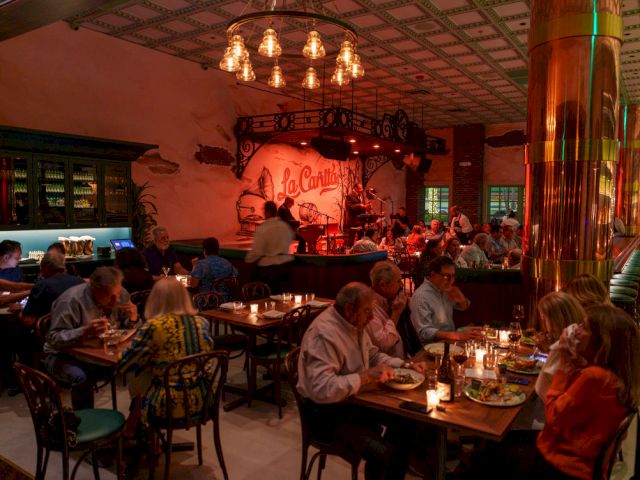 A cozy restaurant with warm lighting, people dining at tables, a live band on stage in the back, and a festive, intimate vibe.