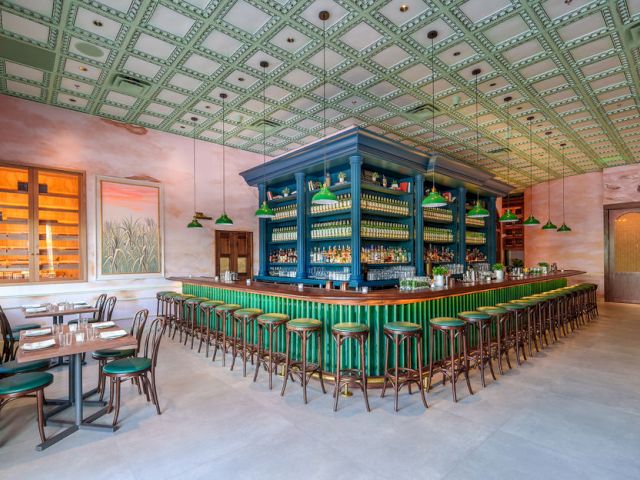A stylish, airy bar with a central blue self-serve counter, green bar stools, and vintage seating; pastel walls and a patterned green ceiling.