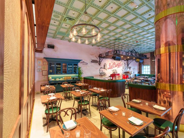 A cozy restaurant with green chairs, wooden tables set with plates, a blue cabinet, warm lighting, and a bar area under decorative ceiling tiles.