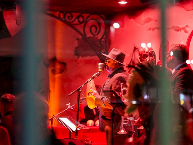 A band performing on stage in a warmly lit venue, seen through a lattice or window foreground, with musicians in hats.