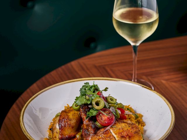 A plated dish with greens and meat on a wooden table, plus a glass of white wine in the background, elegant dining setup.