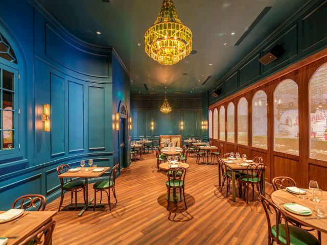 A chic, blue-walled restaurant with warm wood floors, round tables, green chairs, and elegant chandeliers lighting a cozy dining space.