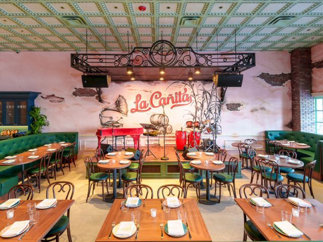 A stylish restaurant with a stage, faux mural &ldquo;La Cantina,&rdquo; vintage chairs, wooden tables, green booths, and a cozy, warm ambiance.