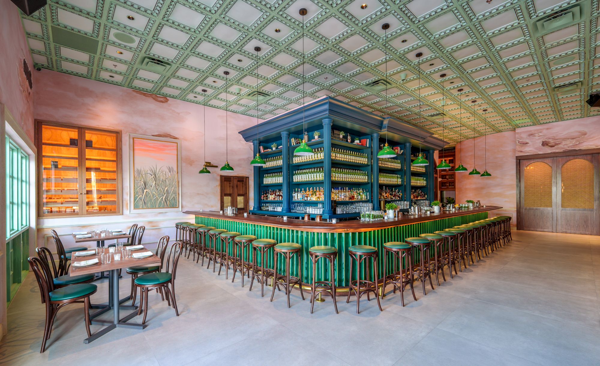 A vintage, airy cafe with a central blue-bar island, green stools, surrounding tables, pastel walls, and a decorative tin ceiling all around.