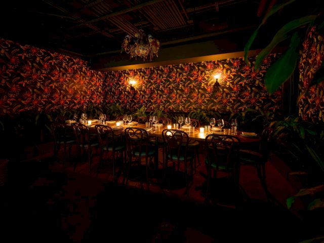 A dimly lit, intimate dining room with floral wallpaper, softly glowing candles on tables, and lush plants creating a cozy, moody atmosphere.