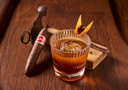 A whisky cocktail in a rocks glass with ice, orange garnish, and a cigar on a wooden bar alongside tools.