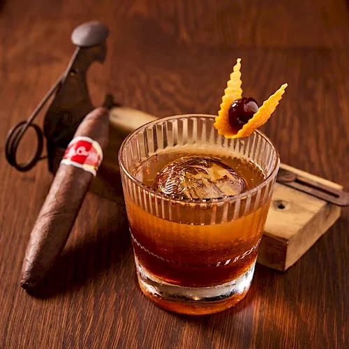 A whisky cocktail in a rocks glass with ice, orange garnish, and a cigar on a wooden bar alongside tools.