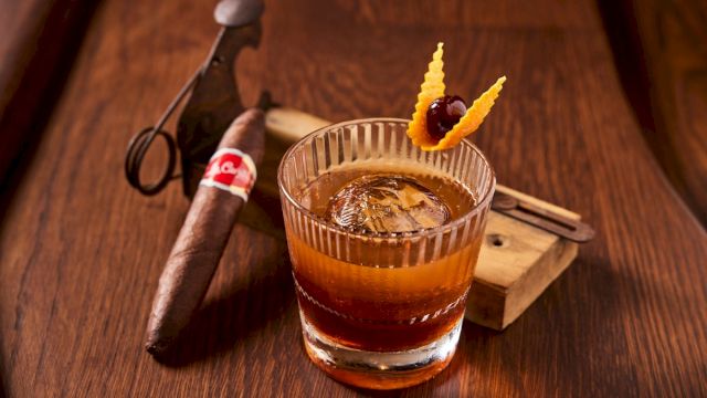 A whisky cocktail in a rocks glass with ice, orange garnish, and a cigar on a wooden bar alongside tools.