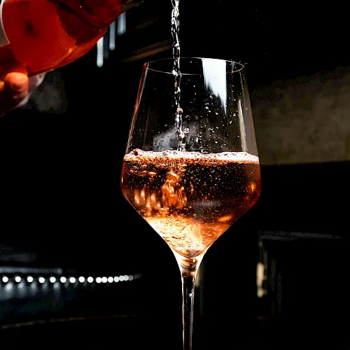 A bartender pours amber wine into a stemmed glass, sparkling as it fills under moody bar lighting.