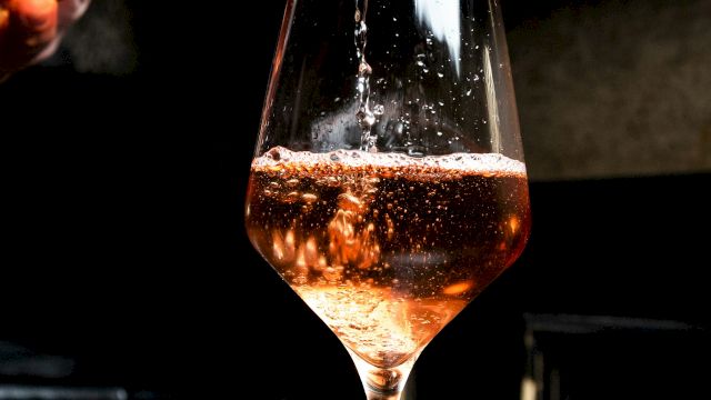 A bartender pours amber wine into a stemmed glass, sparkling as it fills under moody bar lighting.