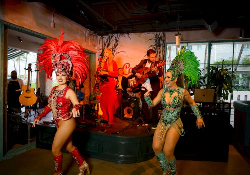 A live band plays as two dancers in bright red and green feather costumes pose onstage at a party, smiling.
