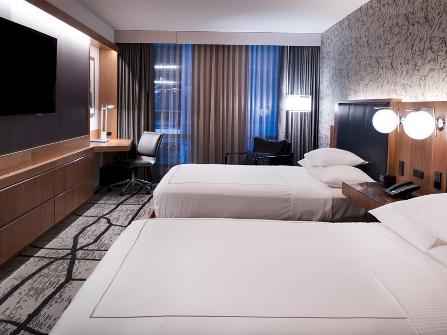 A modern hotel room with two beds, wood paneling, dark curtains, and bright lamps by a window, all neatly made.