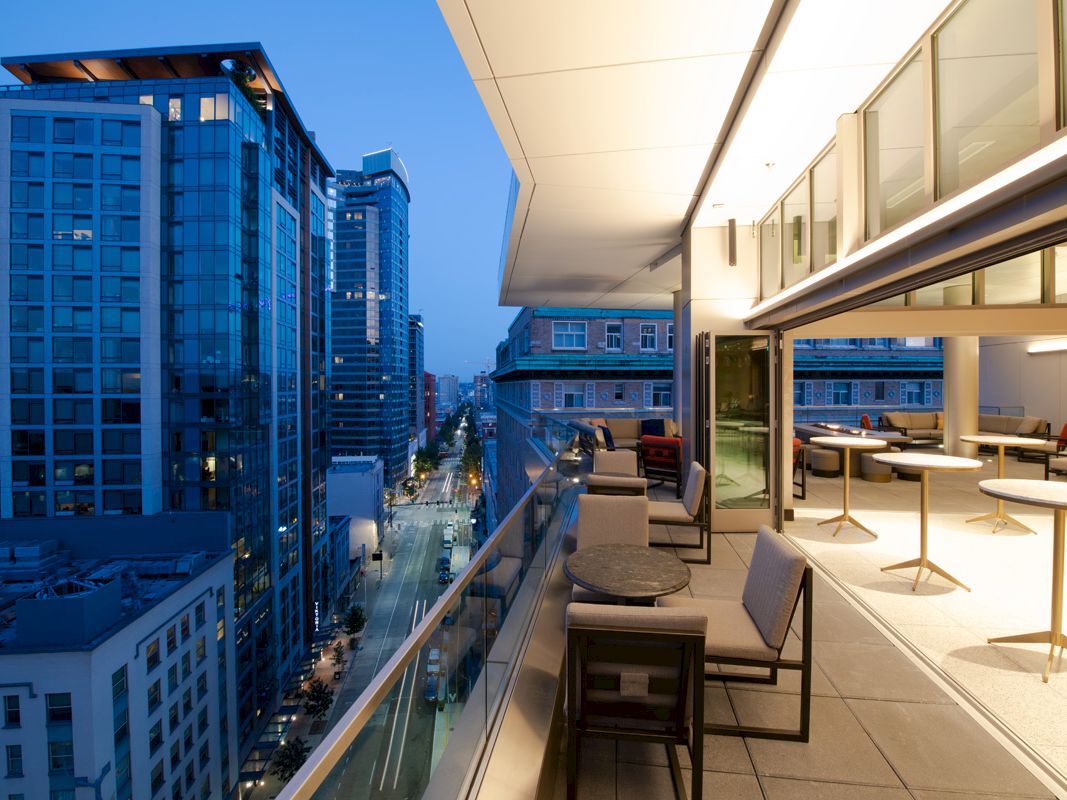 Balcony rooftop lounge overlooking a city street between modern skyscrapers at dusk.